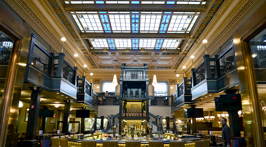 A two story restaurant with a bar in the centre of the room, underneath large skylights and detailed ceilings.