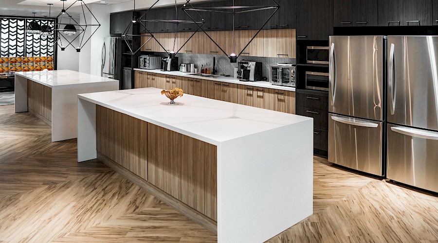 A kitchen with two large white stone islands, a stainless steel fridge, modern light fixtures and wood flooring and panelling.