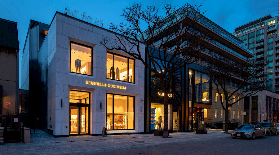 A road in front of the exterior of the Yorkville Avenue retail building occupied by designer stores that are lit up inside in the evening.