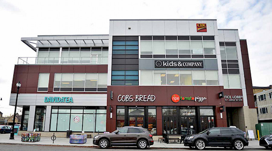 An exterior shot of the Garrison Corner building occupied by multiple retailers, with cars parked along the street in front of the building.