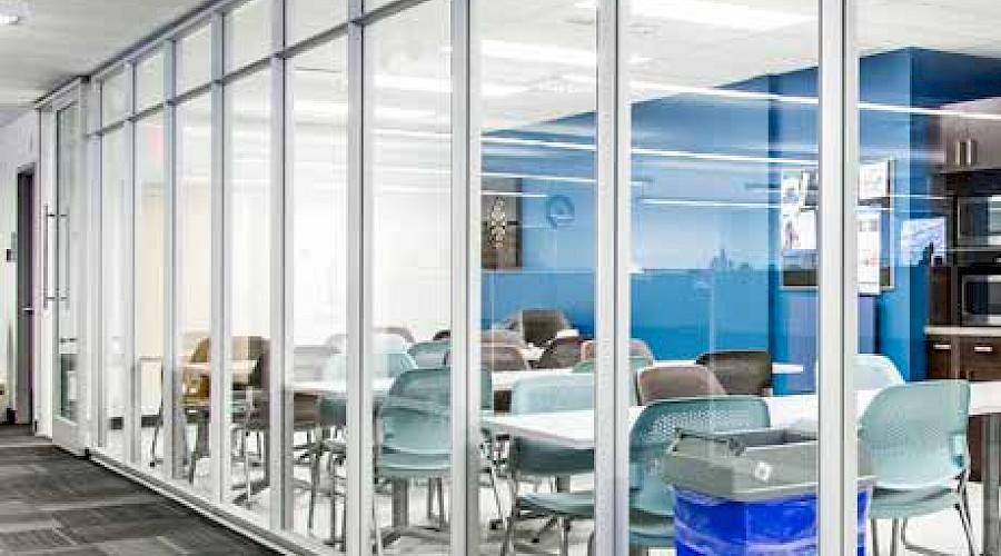 A hallway looking through floor-to-ceiling windows into a classroom filled with desks and chairs.