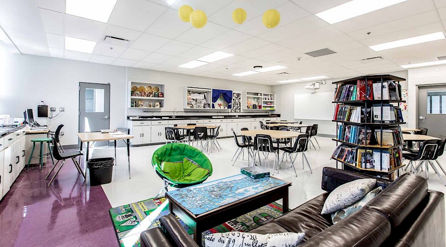 A sofa in a corner of a classroom filled with tables, chairs and a full bookcase.