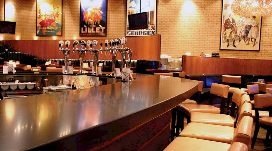 A bar top lined with chairs facing beer taps with large prints on the wall in the background.