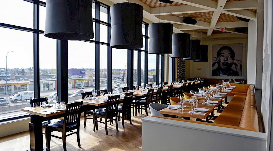 Two rows of tables underneath large black lights fixtures in a second-story restaurant with large windows and a pop culture art print on the far back wall at the Double Zero Pizza in Calgary, Alberta.