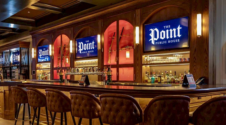 A long bar top facing beer taps and shelves of liquor with TVs on the wall that read 