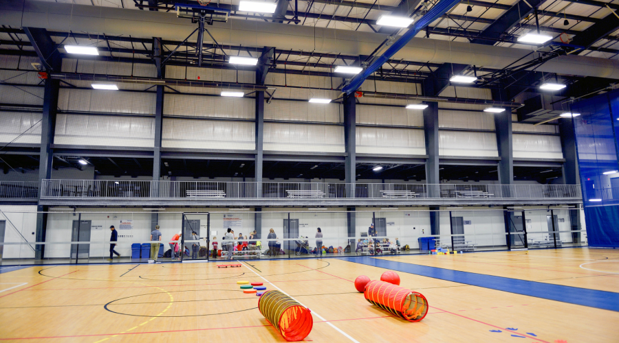 Gym equipment laying on the floor of a large gymnasium with people talking, standing and sitting on the bleachers in the background.