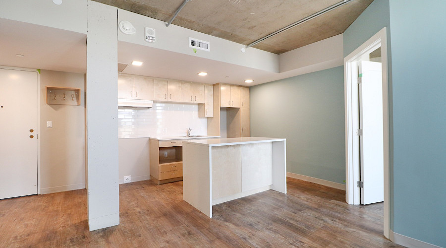 Inside an apartment with wood flooring, white cabinets and appliances and pot lights in the kitchen.