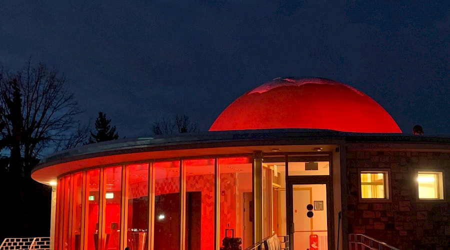 A photo of the Queen Elizabeth II Planetarium lit up red at night against a navy blue sky.