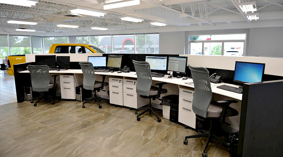 Four chairs behind a large desk with computers and a bright yellow Dodge Ram in the background.