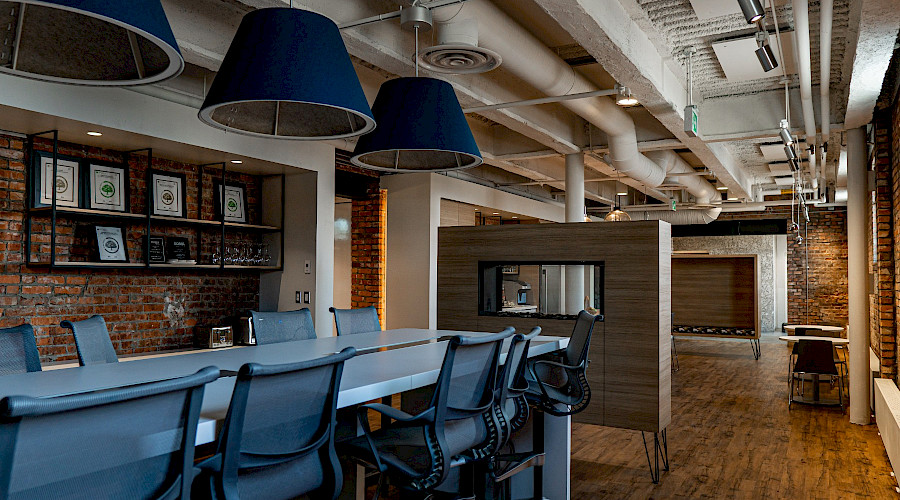 A long desk with multiple chairs inside a room with hardwood flooring and brick walls.