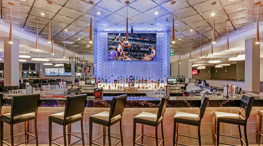 A bar top with multiple open chairs facing a large TV hung above rows of bottles of liquor.
