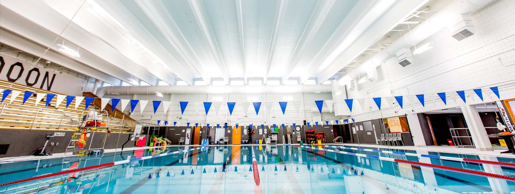 A photo of backstroke flags hanging above a large, bright blue pool under a bright white lit up ceiling.