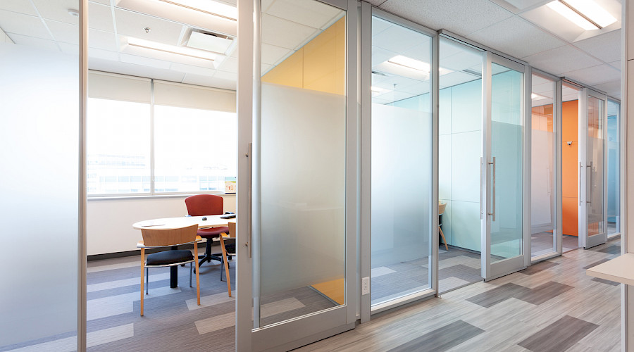 A desk with chairs in a room with frosted glass doors and walls beside other similar rooms down a hallway.