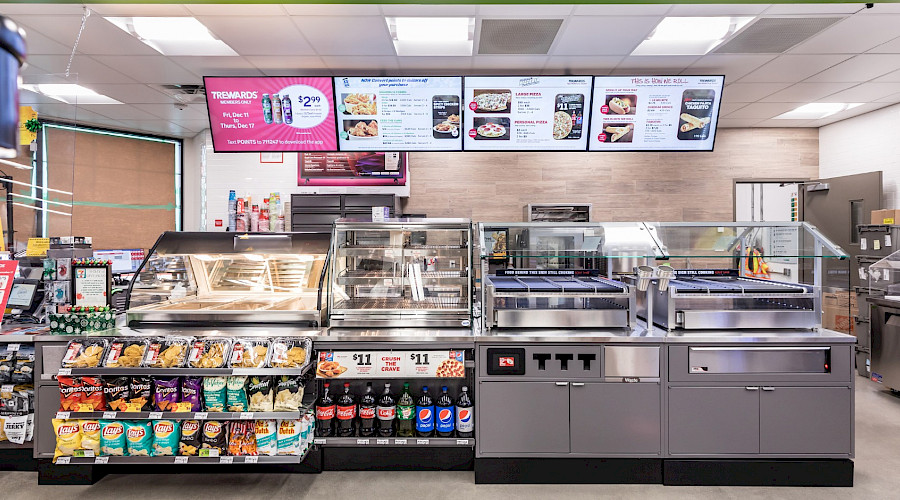 Rows of chips and pop underneath ovens with TVs displaying the menu inside a 7-Eleven.