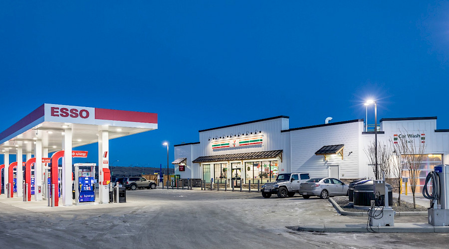 The exterior of 7-Eleven lit up in the evening and the Esso gas pumps in the parking lot.