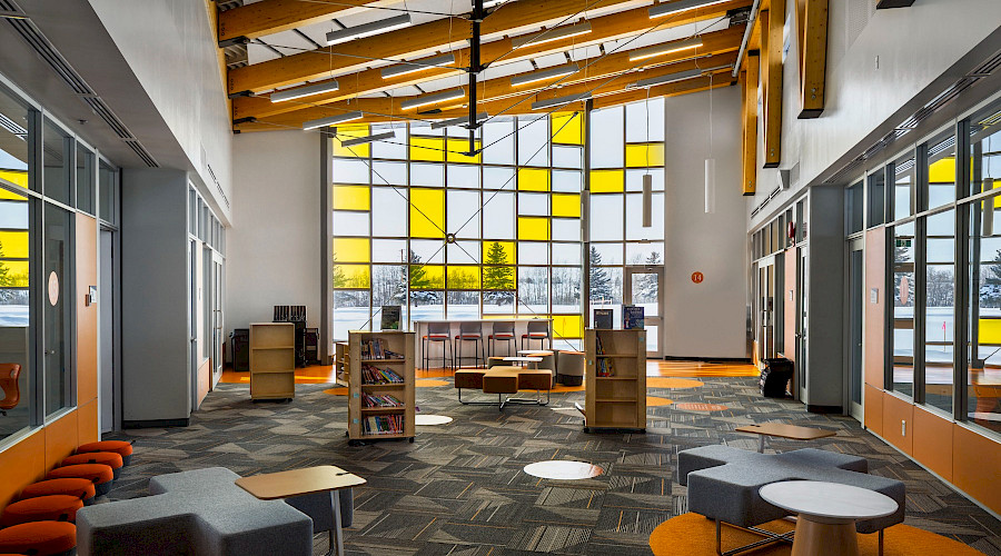 A library with large clear and yellow windows, wooden beams on the ceiling, and orange furniture.