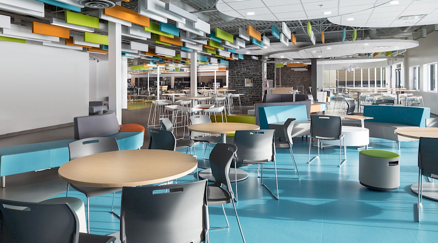 Tables with chairs in a large cafeteria, featuring bright blue flooring and multicoloured panels hanging from the ceiling.