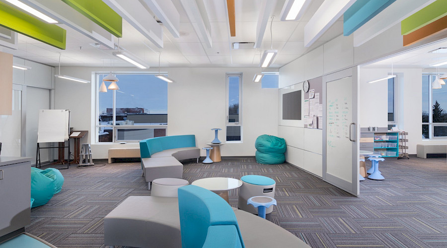 Blue and grey lounge chairs in a room with a bulletin board and a white board and multicoloured panels hanging from the ceiling.