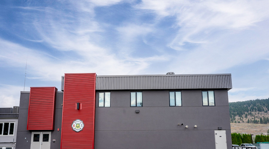 The grey and red exterior of the Merritt Firehall behind a patch of green grass.