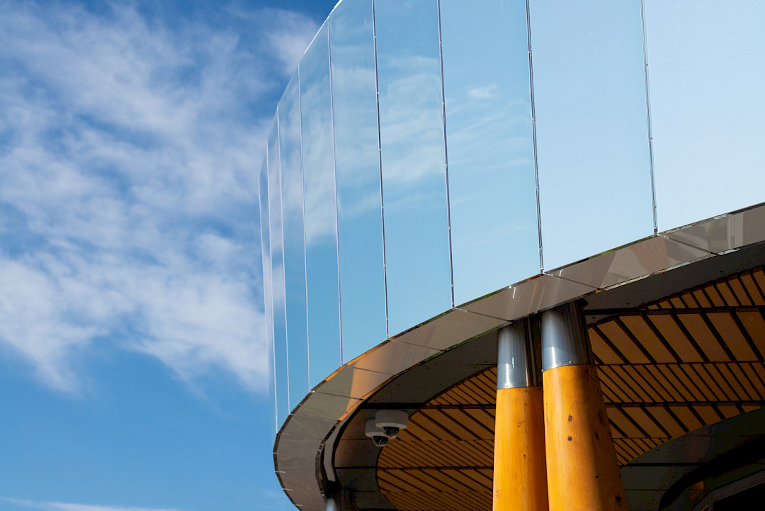 A photo of a rounded corner of the Blatchford energy centre building with a mirrored building envelope against a blue sky with clouds.