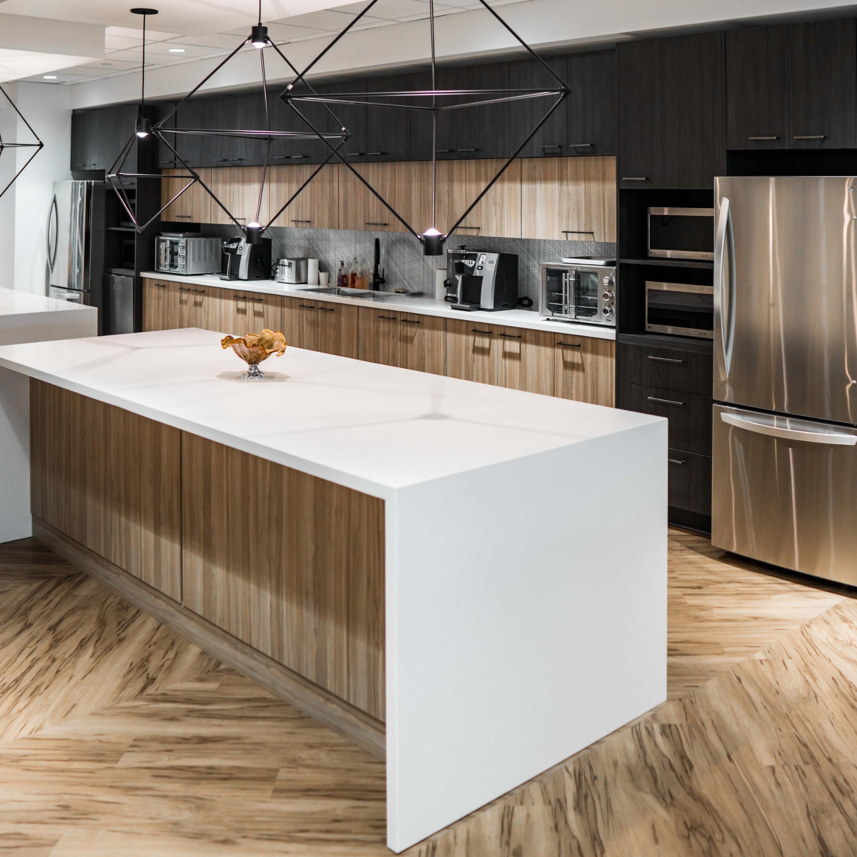 A kitchen with a large white marble island, modern black light fixtures, stainless steel appliances and light hardwood floors and wooden cabinets.