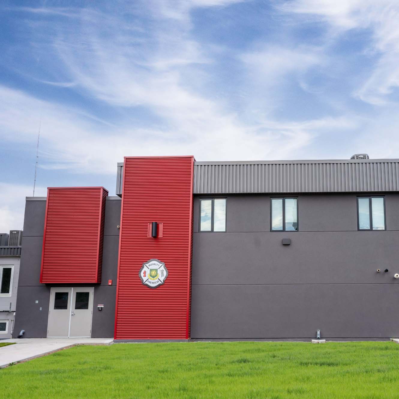 The red and grey exterior of the Merritt Firehall with a cloudy blue sky in the background.