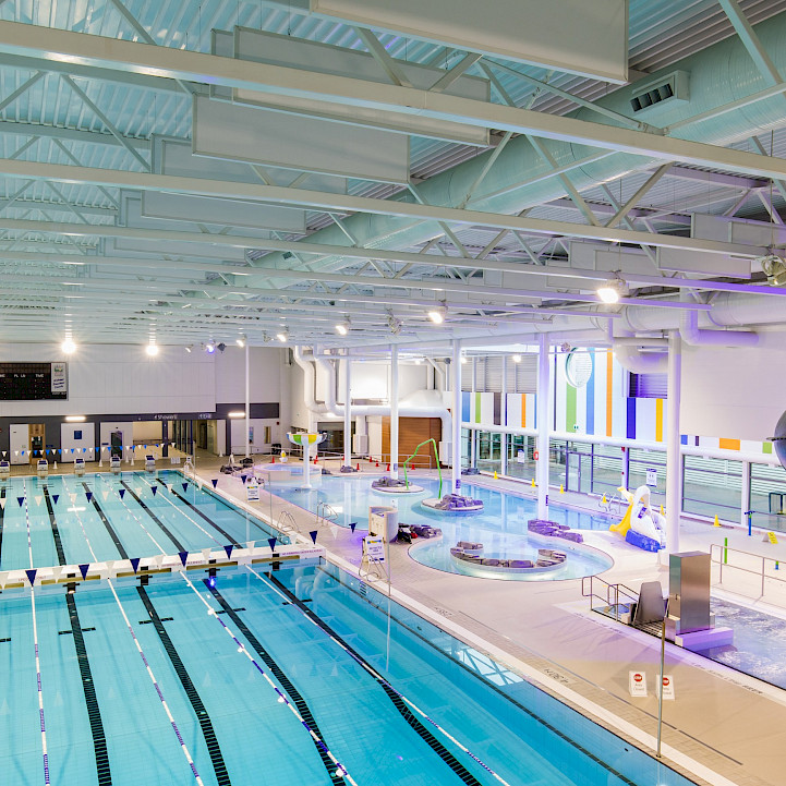 An elevated shot of the Canada Games Aquatic Centre pool, featuring five swimming lanes, a lazy river, and bright lights.