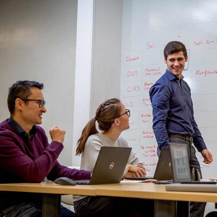 Two Chandos employees sitting at a desk with their computers, taking notes while they discuss work with another Chandos employee using the whiteboard to express ideas.