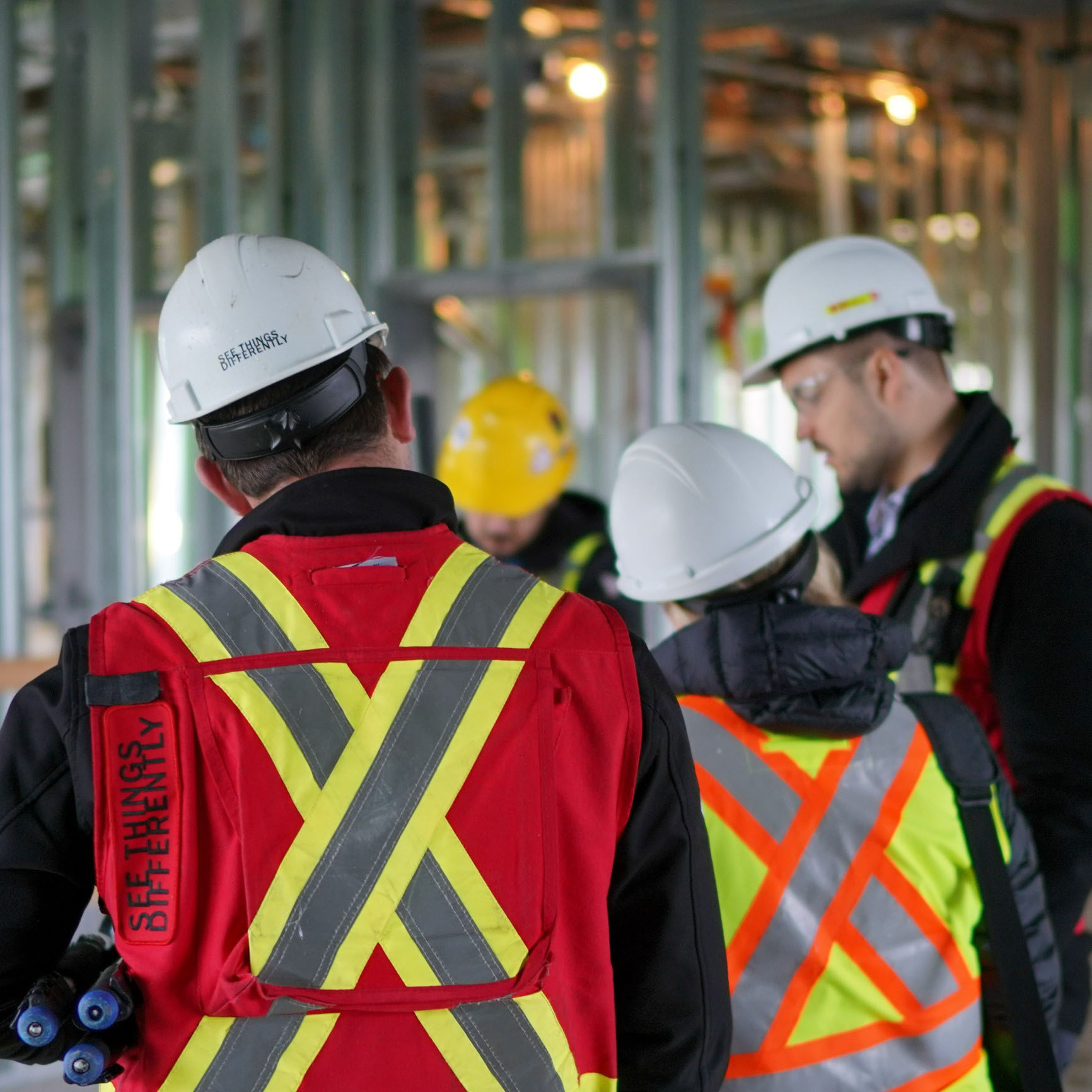 Three Chandos construction workers facing away from the camera, discussing a site on project, wearing safety vests and hard hats that read 