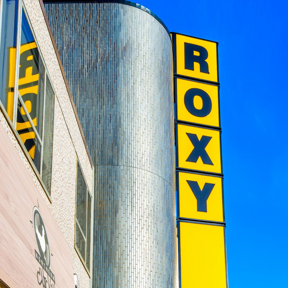 A photo of the exterior of the Roxy Theatre, featuring it's large windows and bright yellow vertical sign that reads 