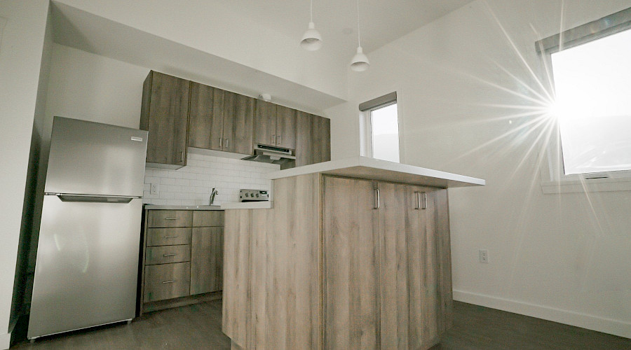 The sun shining into a kitchen with a small island, wooden cupboards and a stainless steel fridge.