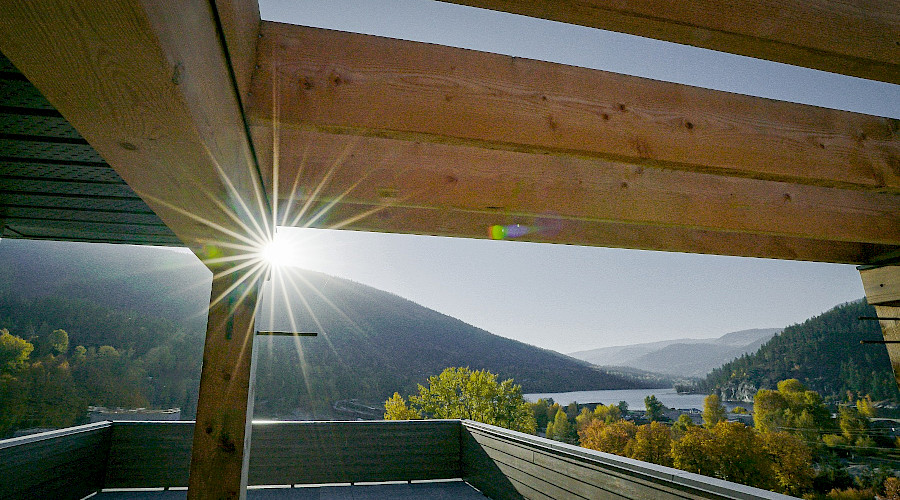 The sun shining onto the covered wood roof of a building, with orange and green trees, a lake, and hills in the background.