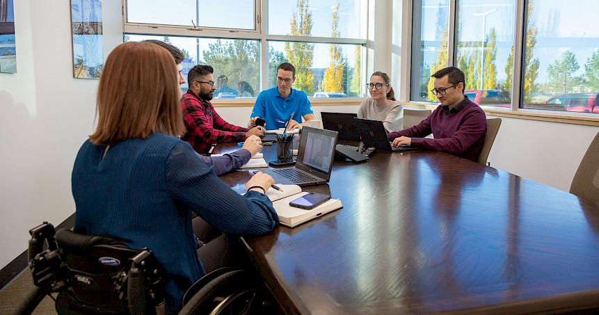 A group of diverse Chandos construction office workers sitting at a table in bright room, all working on their computers and discussing.
