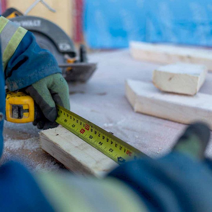 Two gloved hands using a measuring tape to measure a piece of wood on a construction site.