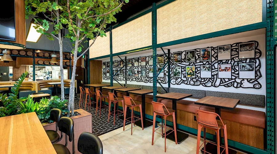 A row of high top tables along a wall lined with pictures across trees and plants. At the The Central Taps and 33 Acres restaurant and 33 Acres Brewing bar in downtown Calgary