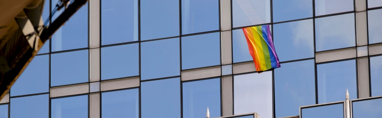 A photo of a rainbow flag waving in front of a building with glass windows.