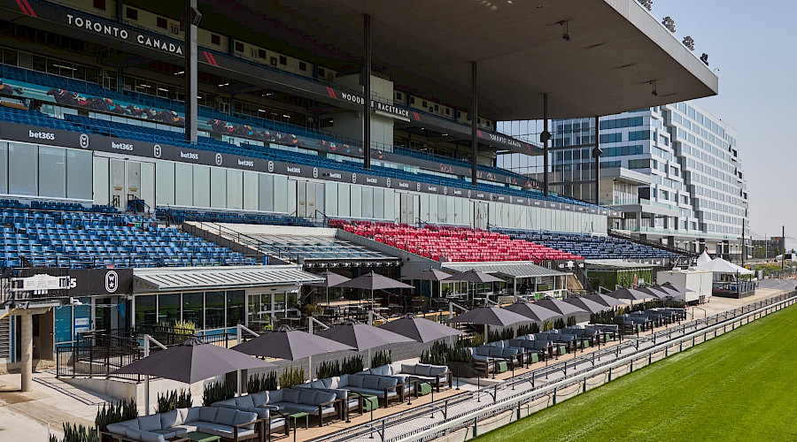 Drone image of the new Woodbine Apron Patio built by Chandos Construction Toronto. The image features a long row of patio tables shaded by grey umbrellas, the outdoor kitchen, bar seating with custom wood table tops, and it showcases how close the seats are to the race track.