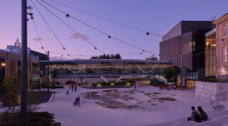 Exterior image of the new courtyard at the Aurora Town Square centre, built by Chandos Construction Toronto. The courtyard is lit at dusk and features community members enjoying the space in the summer.